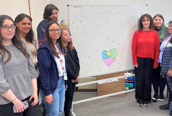 A group of young people and adults stand around a whiteboard in a classroom or meeting room. The whiteboard is decorated with colourful confetti-style markings and reads “Welcome to Moray” with a multicoloured heart drawn underneath. Posters are displayed on the walls, and the group faces the camera beside the welcome display.